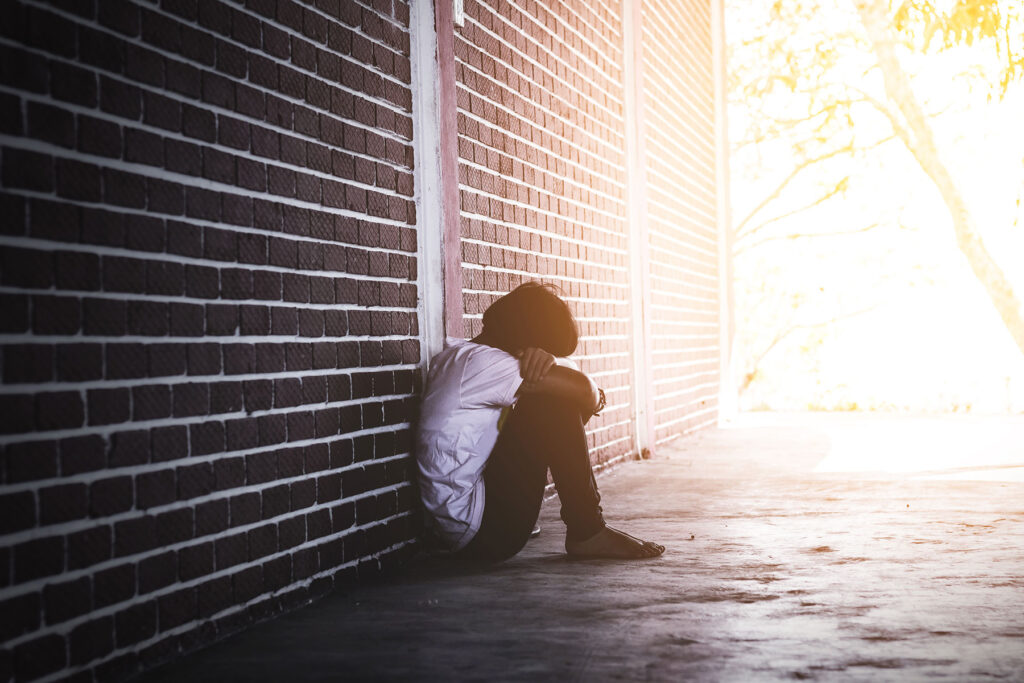 Depressed teen sitting outside against a wall with his head down while struggling with signs of trauma in adolescence