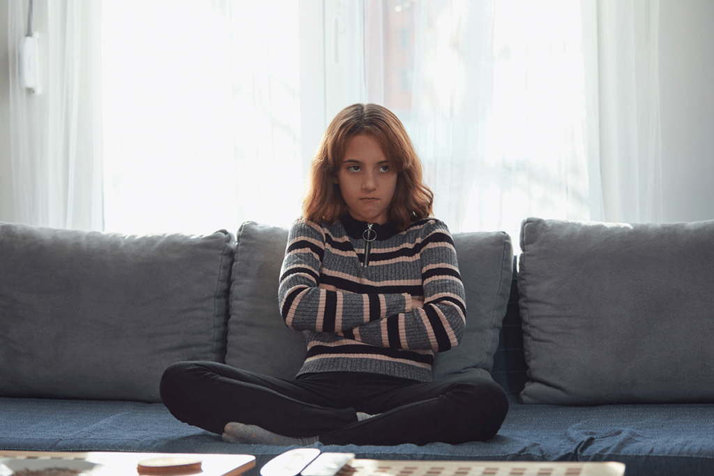 Adolescent sitting on couch with arms crossed and stern look on her face struggling with common grief responses